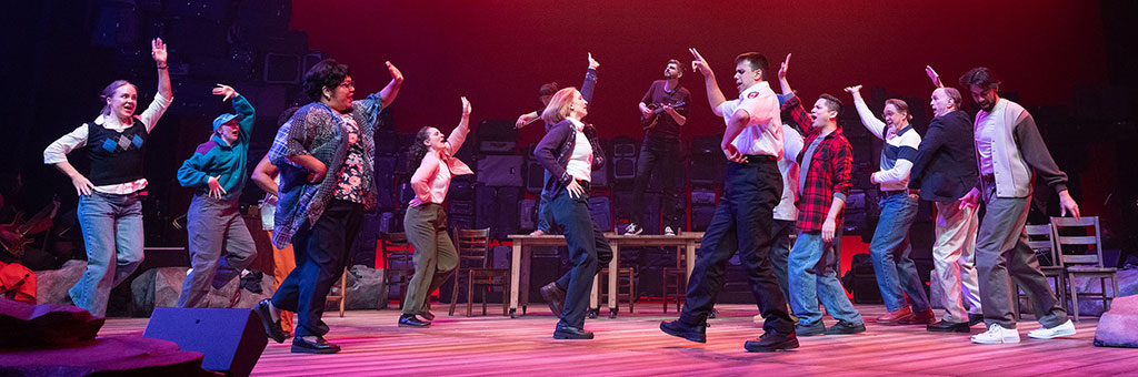 Group of performers dancing together on a stage under red and blue lighting with raised arms and energetic movement.