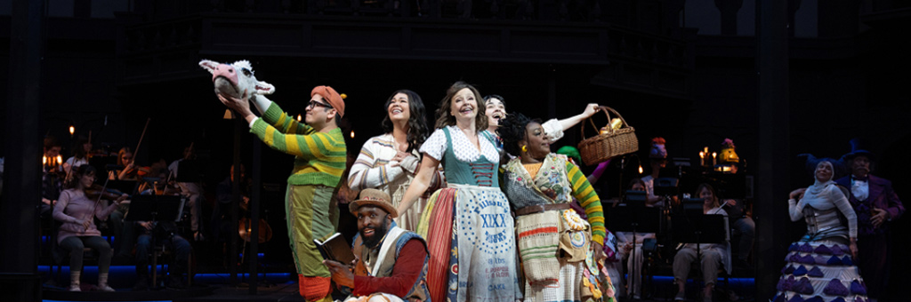 Ensemble cast on stage, some holding props, with orchestra in background.