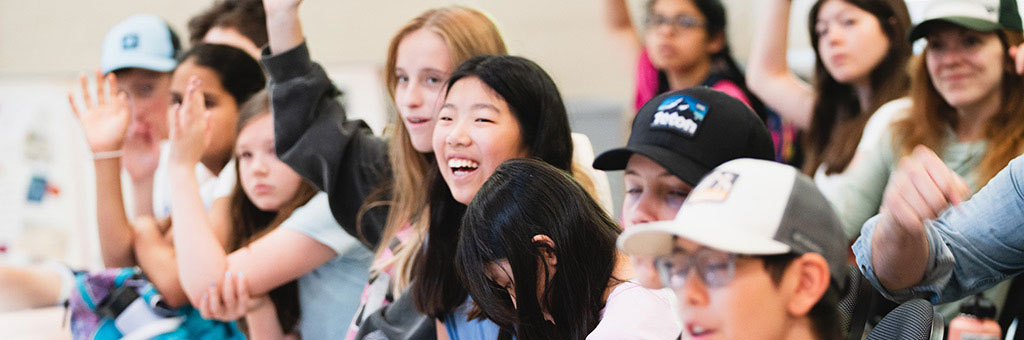 A group of individuals sitting in a classroom-like setting, some raising their hands enthusiastically. They appear engaged and attentive, with a mix of expressions including smiles and curiosity.