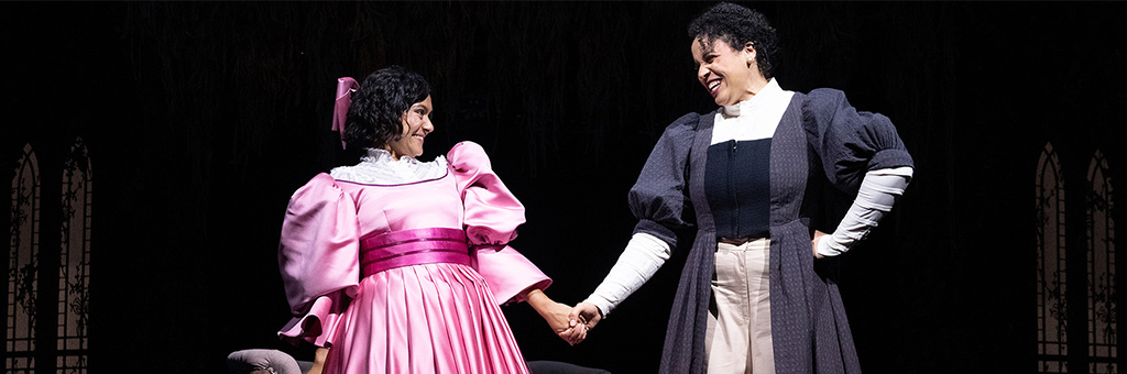 Two people on stage hold hands and smile at each other. One wears a bright pink, voluminous dress with a large bow, and the other is dressed in a darker, more subdued outfit with white sleeves and a long skirt. The backdrop suggests an ornate, darkened set with tall, arched windows.