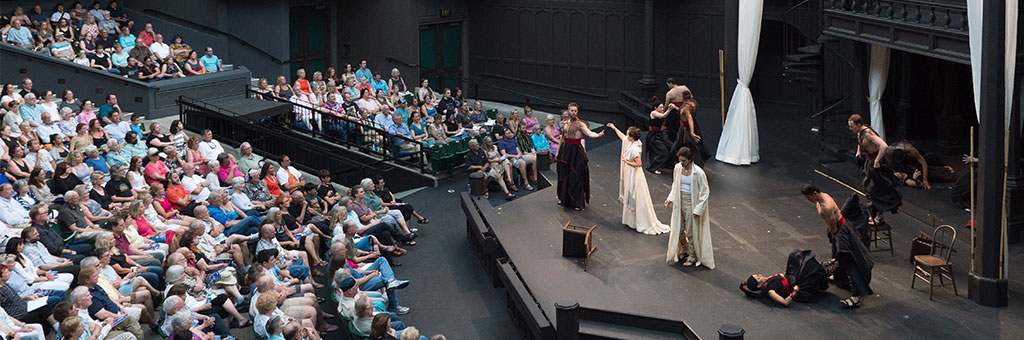 Actors perform a dramatic scene on an outdoor stage at the Oregon Shakespeare Festival, dressed in period costumes with flowing fabrics and dark cloaks. Some stand in intense poses, while others kneel or lie on the stage. A large audience watches from tiered seating in the background.