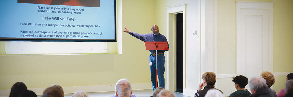 A person stands at a podium in front of a projected slide discussing “Free Will vs. Fate” in Shakespeare’s Macbeth, speaking to an audience in a brightly lit room.
