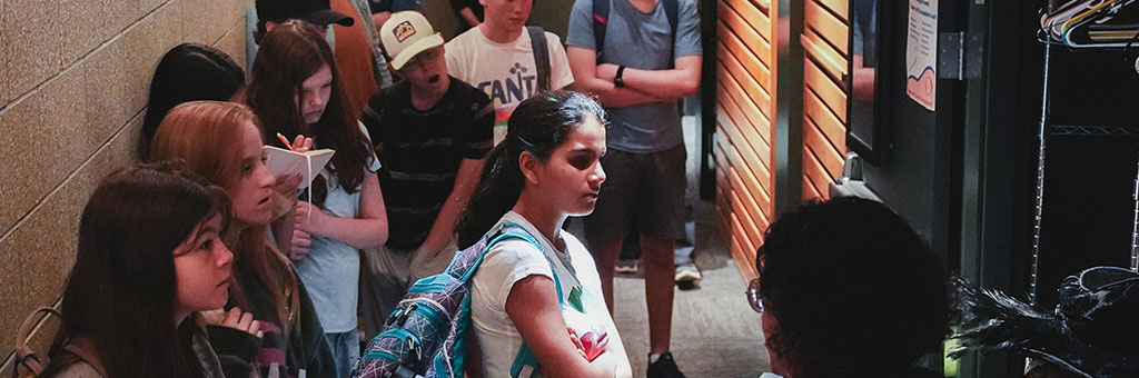 A group of people standing in a narrow hallway, attentively listening to someone at the front. The group appears engaged, some with backpacks and notepads, while looking at an open closet or storage area with hangers.