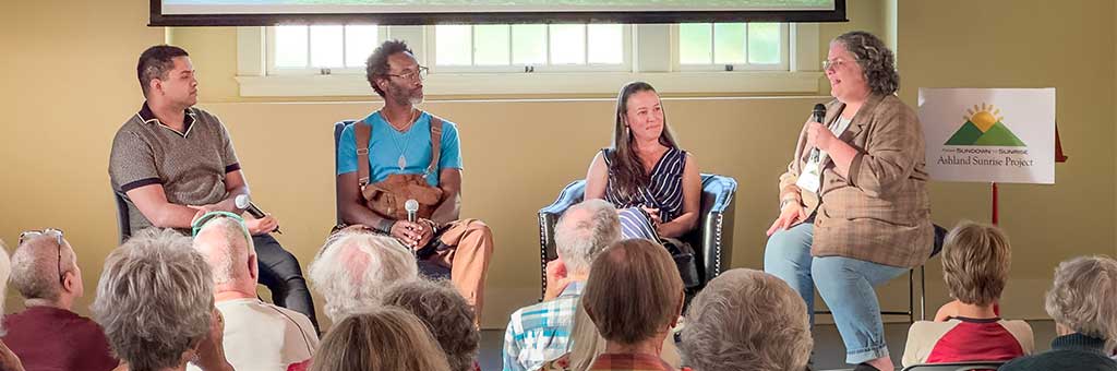 Four panelists sit on stage in front of an audience, each holding microphones. A sign behind them displays "Ashland Sunrise Project," indicating the event theme.