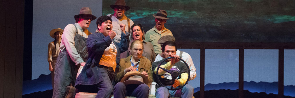 Group of performers crowded in a car prop, singing passionately against a dark backdrop.