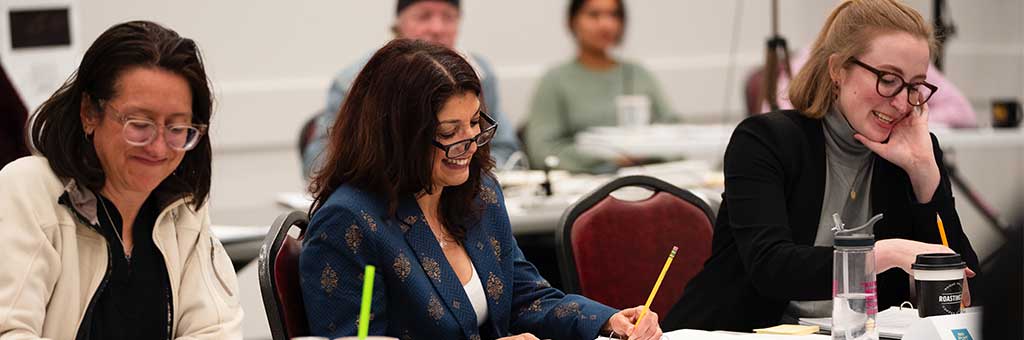 Three people sitting at a table in a rehearsal room, each engaged in writing and smiling. The individuals have different hair colors and are wearing glasses. The background includes blurred figures and various furniture.