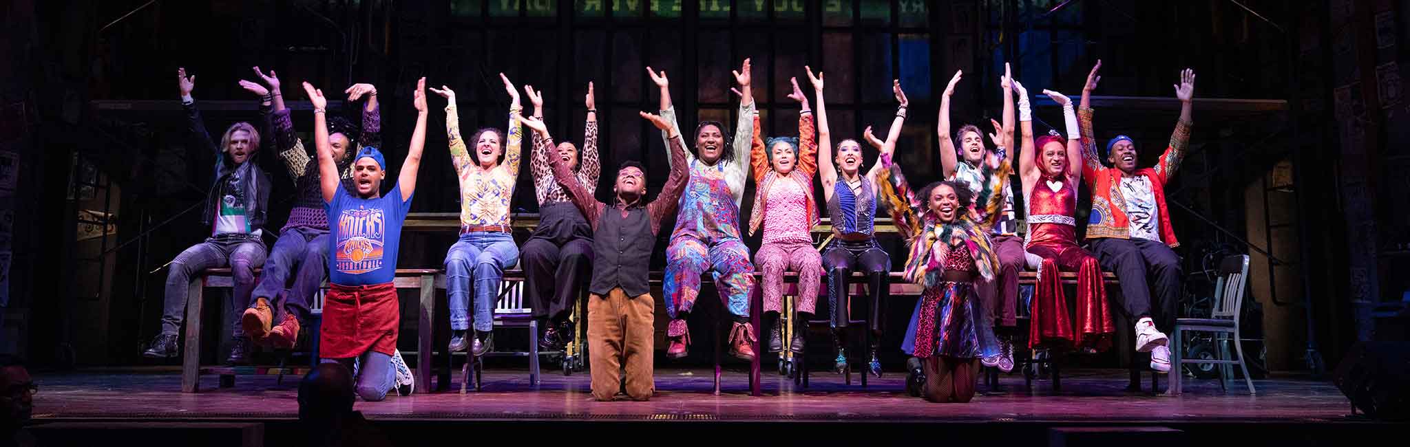 The cast of Rent with their hands in the air singing.