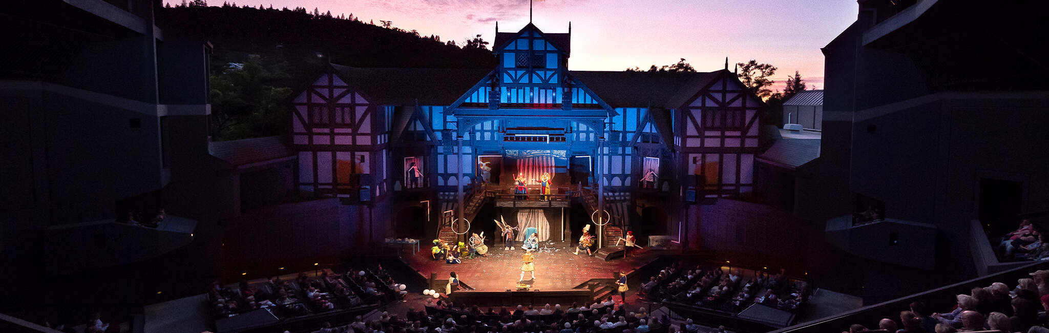 Outdoor theater performance on a multi-level stage styled like a Tudor building at dusk, with actors performing before a seated audience.