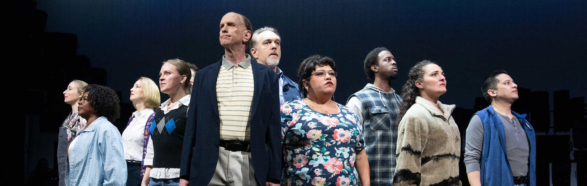 A group of performers stands in a line on a dark stage. Each person faces a different direction with a serious expression.