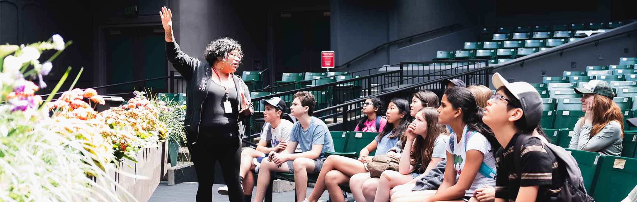 A group of people, mostly young students, sits attentively in an outdoor amphitheater while a guide speaks to them. The guide gestures with one hand, and the audience listens intently.