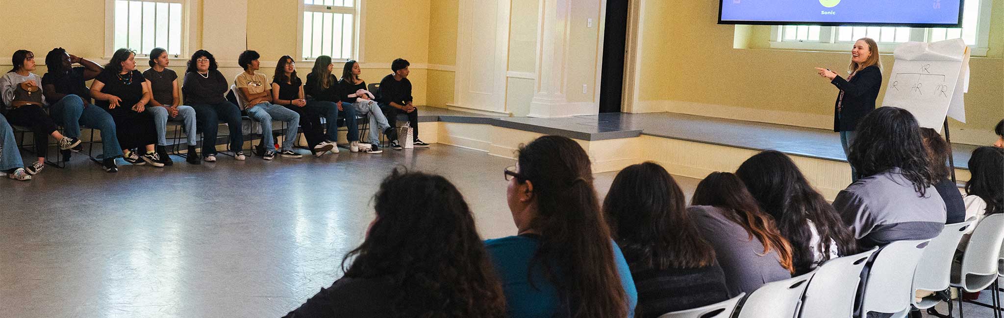 Participants seated in a circle and audience seating area during a group discussion in a community meeting room.