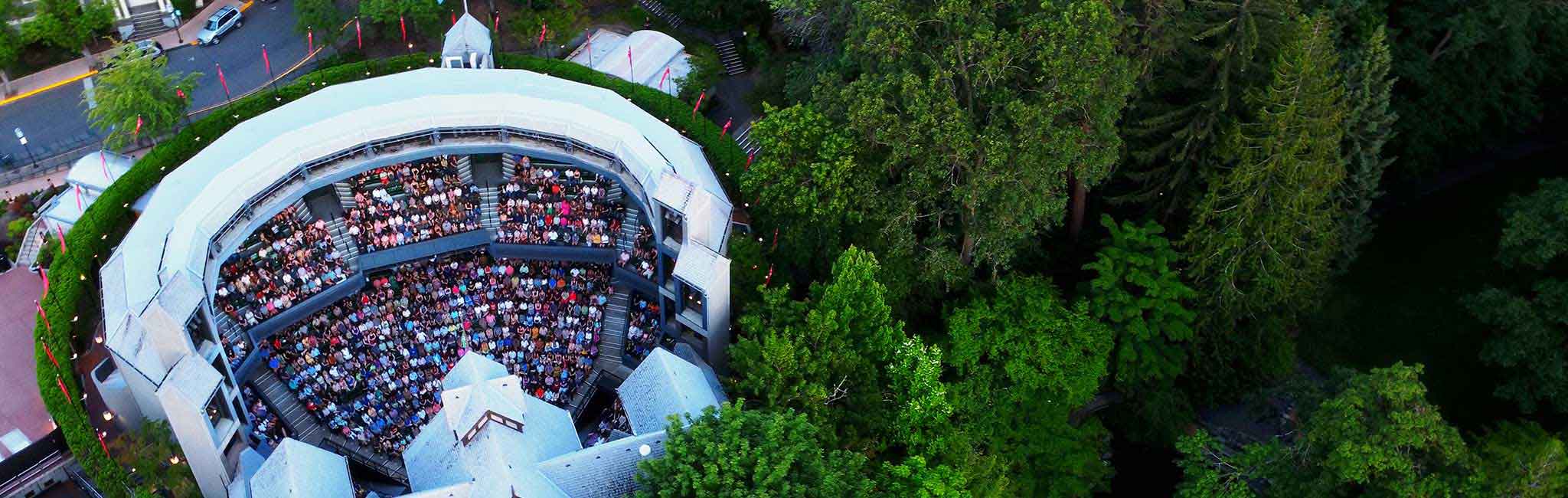Aerial view of the Allen Elizabethan Theatre, showcasing its open-air seating filled with a large audience. The iconic semi-circular structure is surrounded by lush green trees, creating a picturesque setting.