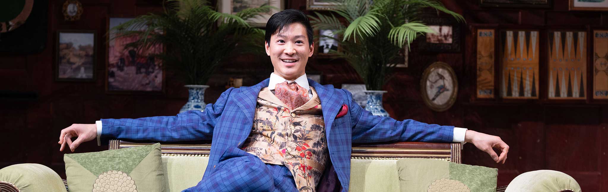 A performer in an ornate blue suit lounges confidently on a green sofa on a stage set decorated with framed art and plants, smiling at the audience.