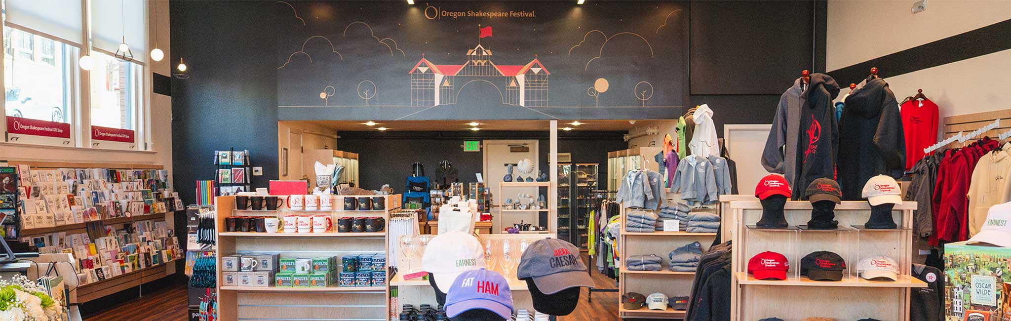 Interior of the Oregon Shakespeare Festival gift shop featuring racks of books, shelves of branded mugs and hats, and displays of hoodies and jackets. A mural of a stylized theater building and trees decorates the back wall above the entrance.