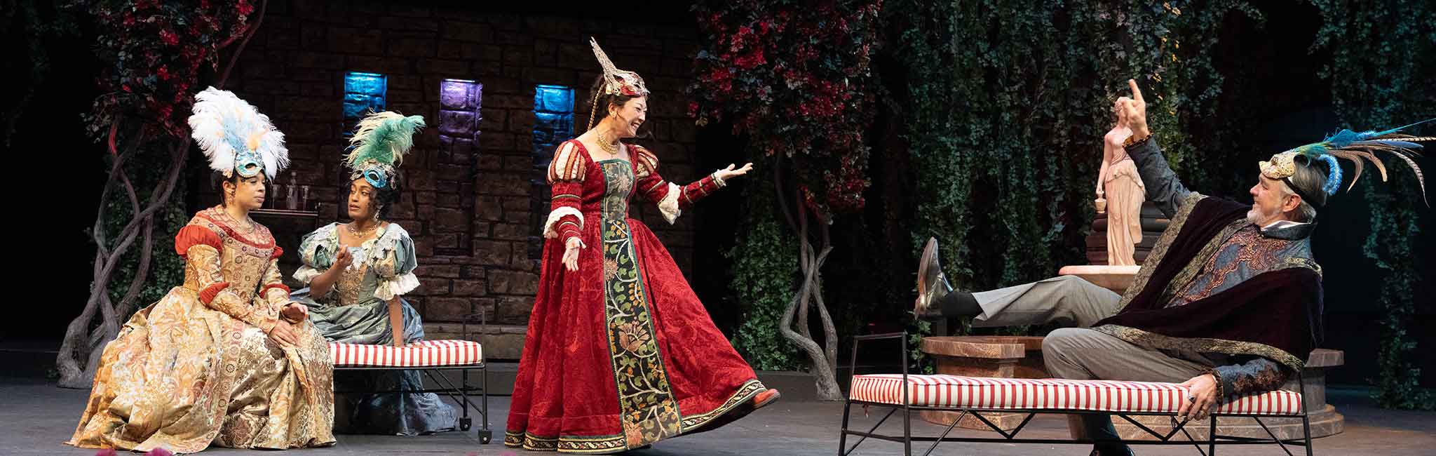 Performers in ornate period costumes and feathered headpieces acting on a stage with a garden-themed backdrop. One person lounges on a striped bench, gesturing dramatically.