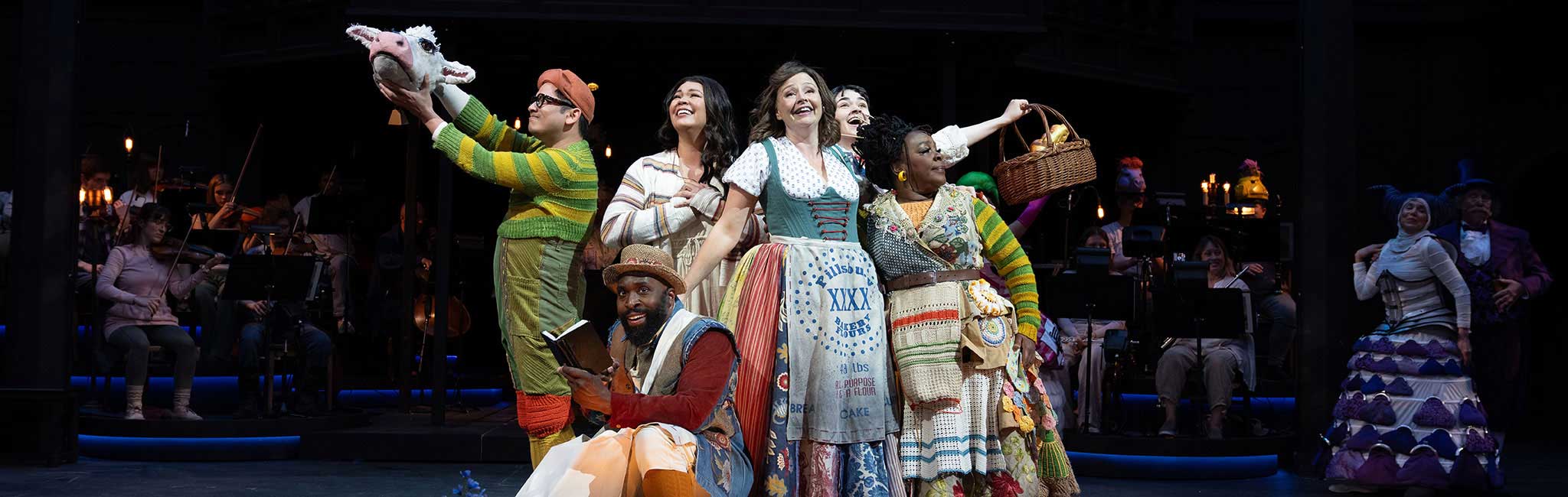 Ensemble cast on stage, some holding props, with orchestra in background.