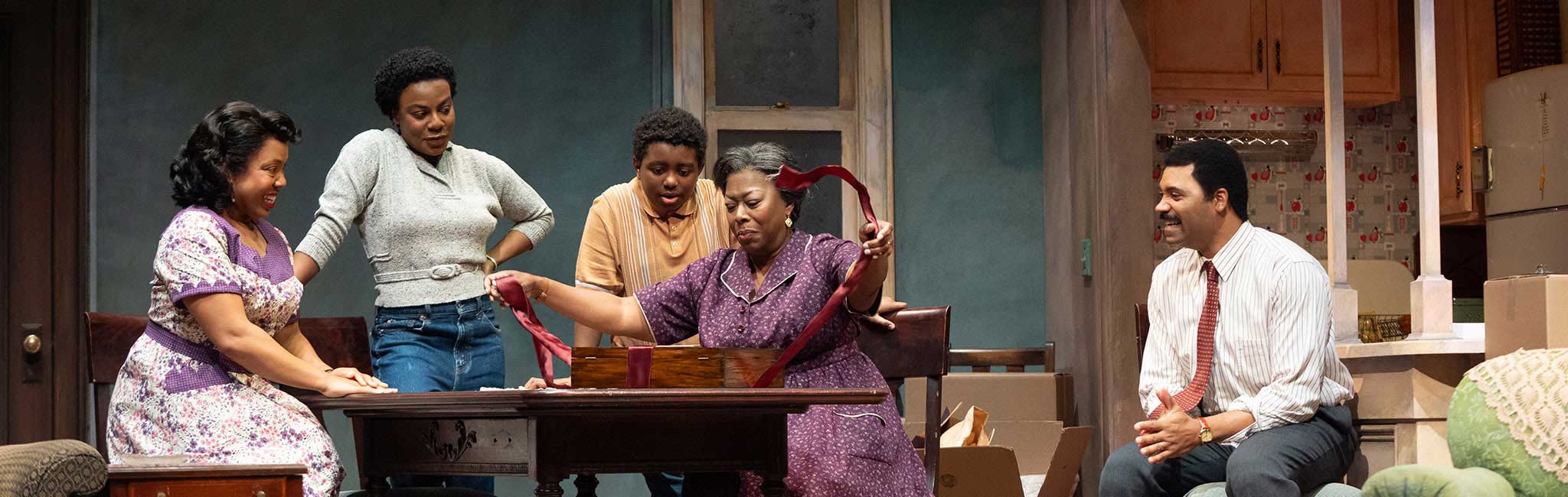 Five people gathered around a dining table on a stage set while one person unwraps a ribbon from a box and the others watch and smile.