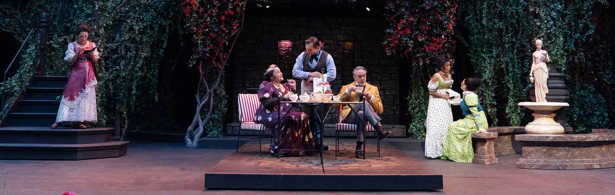 Stage scene set in a lush garden courtyard with actors seated at a small table having tea while others stand nearby under hanging greenery and flowers.