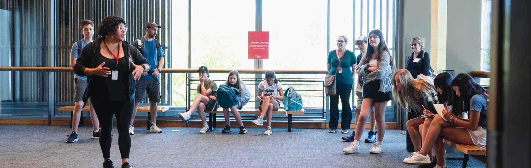 A facilitator addresses a group of students in a bright campus lobby, with some standing and others seated along a bench listening or using notebooks and phones.