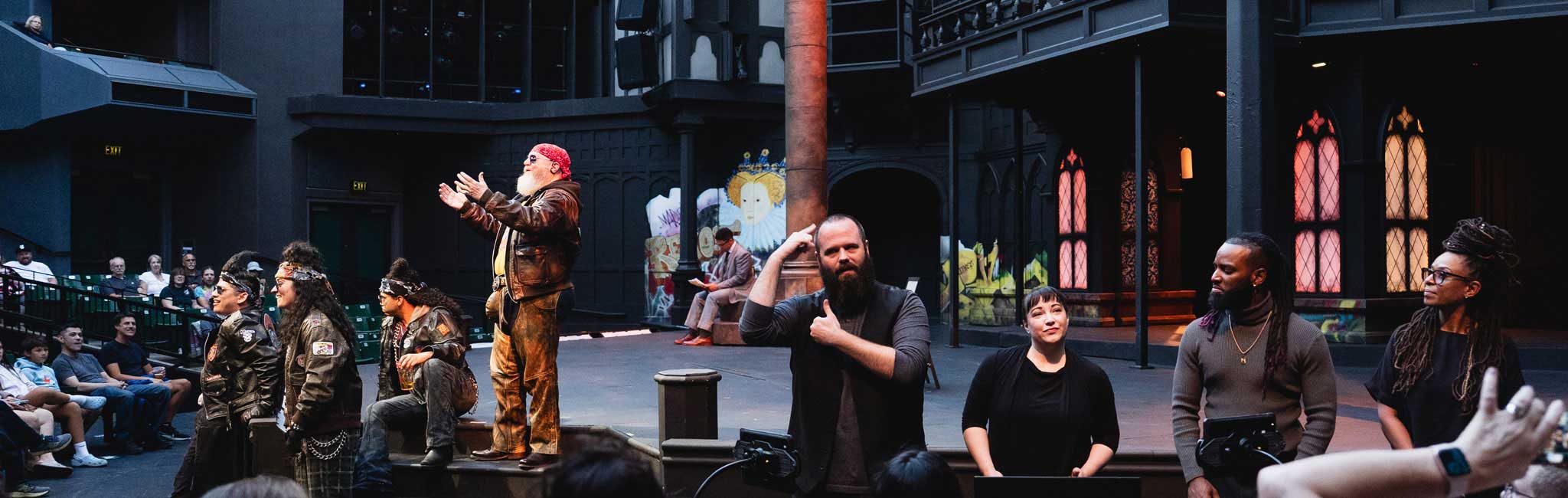ASL interpreters perform at the front of a theater stage while actors continue the play behind them. Audience members watch from nearby seats.