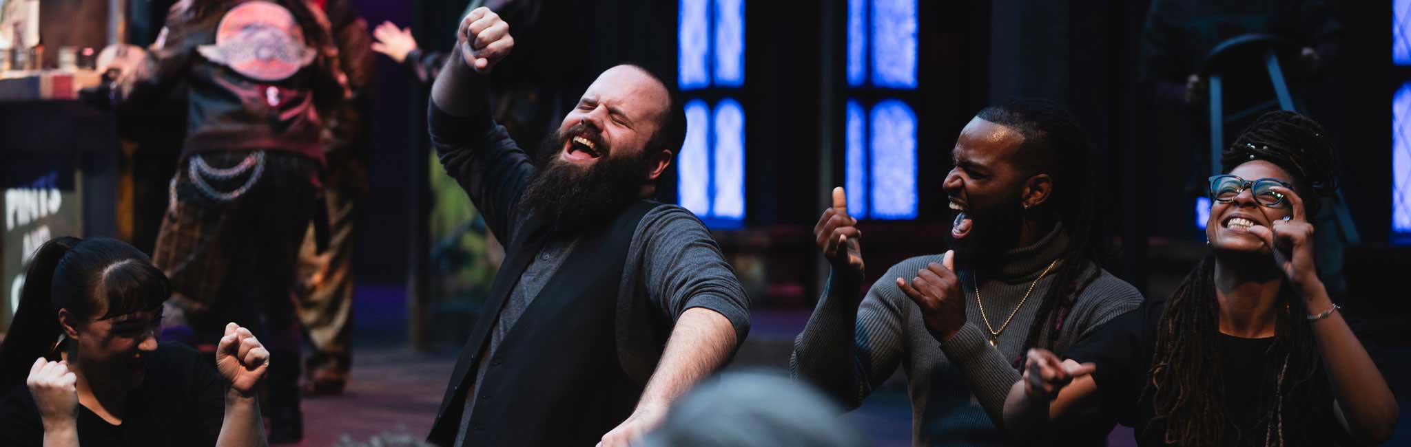 A group of ASL interpreters energetically interpret a play with expressive faces and raised hands. Blue stage windows glow behind them.
