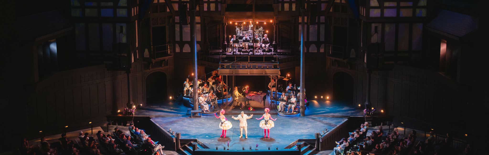 A wide theater view shows three performers at center stage with musicians arranged behind them. Audience members sit on both sides beneath dramatic blue and amber lighting.