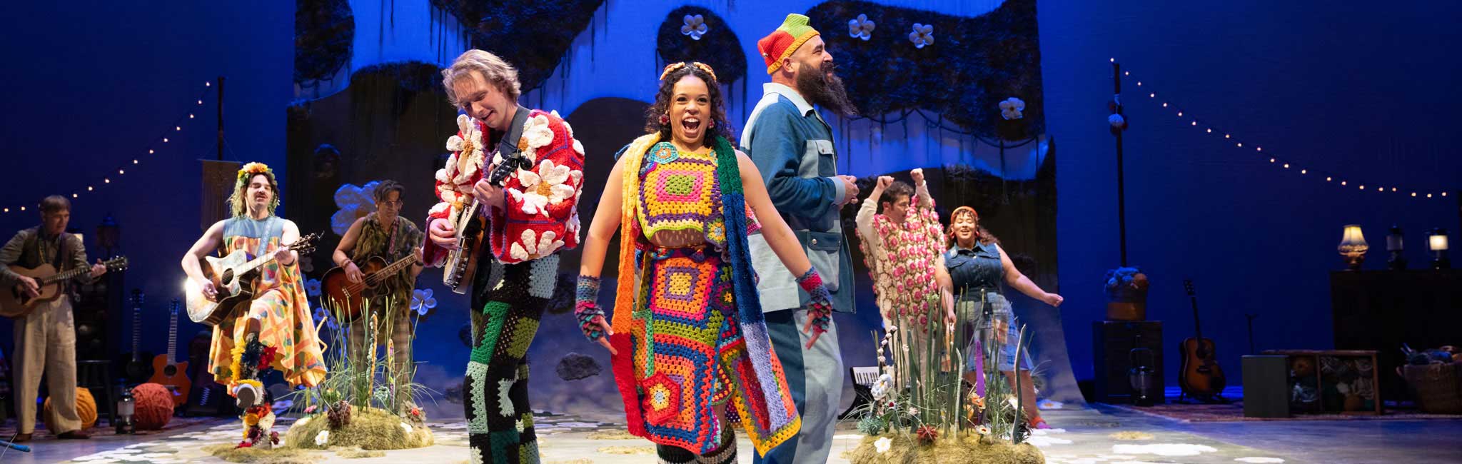 Cast performing with guitars and bright crochet costumes on stage.